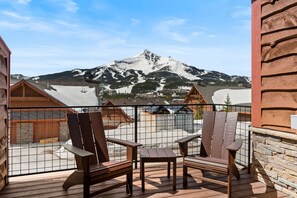 A wooden deck with Adirondack chairs and a table, overlooking a snowy mountain and a ski resort.