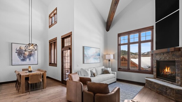 A spacious living room with a fireplace, a dining area with a wooden table and chairs, and large windows offering a view of the outdoors.
