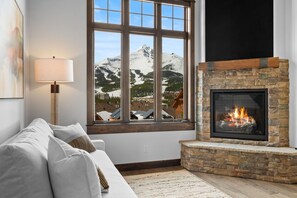 A cozy living room with a stone fireplace, a large window with a mountain view, and a modern lamp.