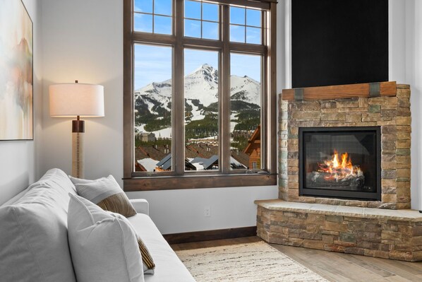 A cozy living room with a stone fireplace, a large window with a mountain view, and a modern lamp.