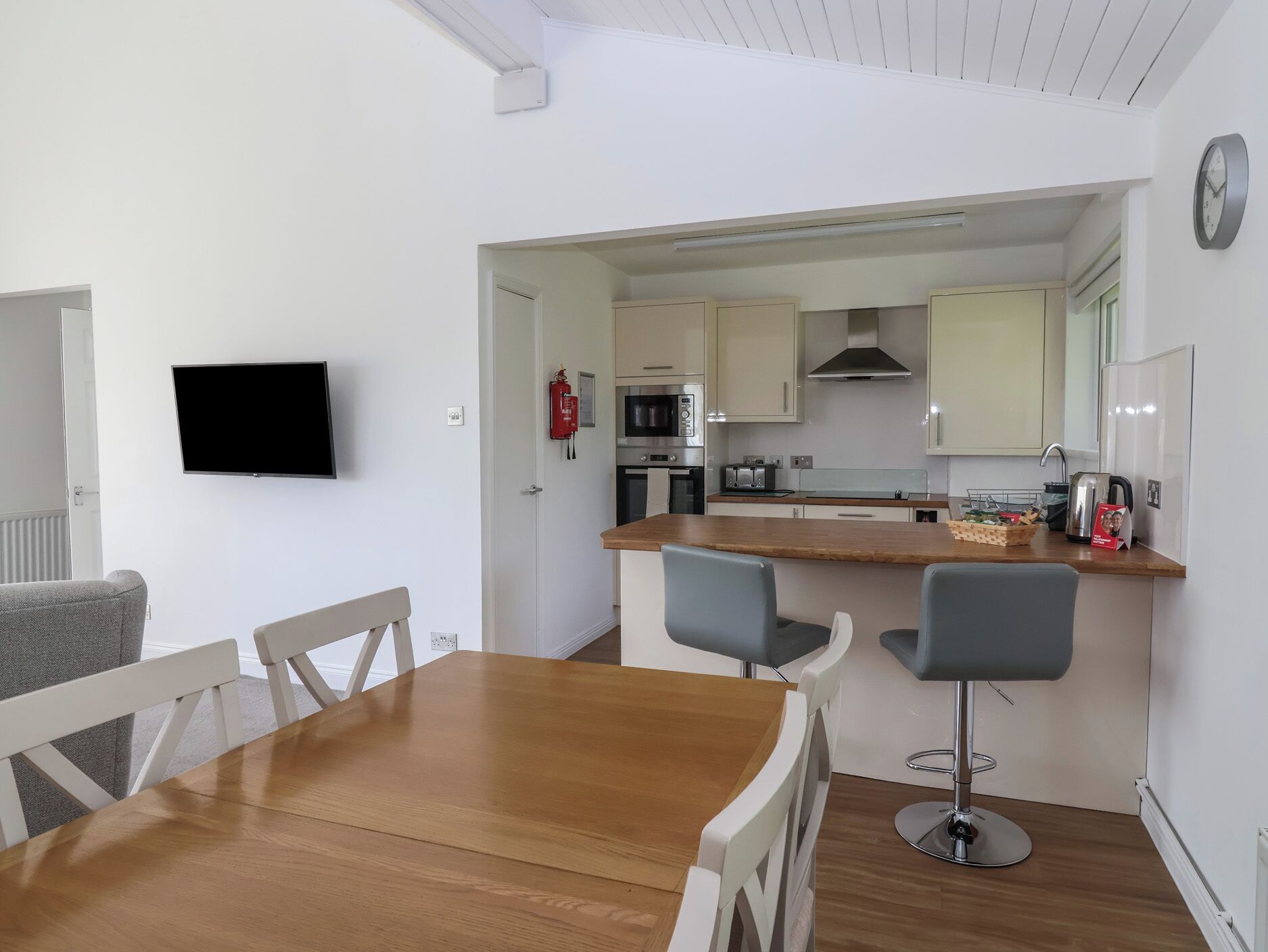 Een moderne keuken met een houten eettafel, twee barkrukken en een aan de muur bevestigde televisie.
