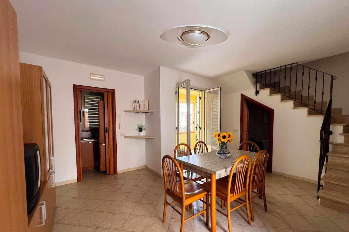 A dining area with a table, chairs, and a vase of sunflowers. There is a staircase to the right and a door leading to another room.