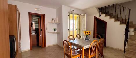 A dining area with a table, chairs, and a vase of sunflowers. There is a staircase to the right and a door leading to another room.