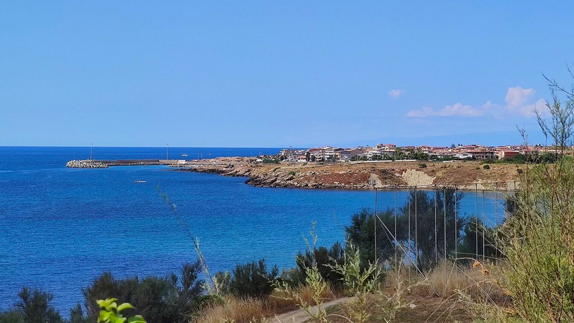 A coastal view with a rocky shoreline, a clear blue sea, and a small settlement with buildings.
