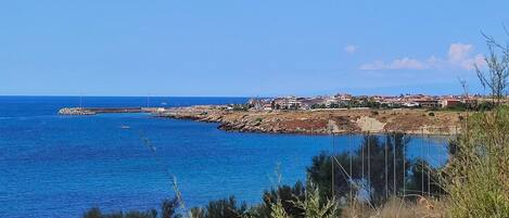 A coastal view with a rocky shoreline, a clear blue sea, and a small settlement with buildings.