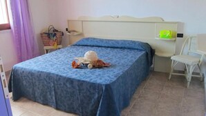 A bedroom with a bed covered in a blue floral bedspread, a wicker basket, and a small chair. - Tucano Apartment Club Resort (Isola di Capo Rizzuto)