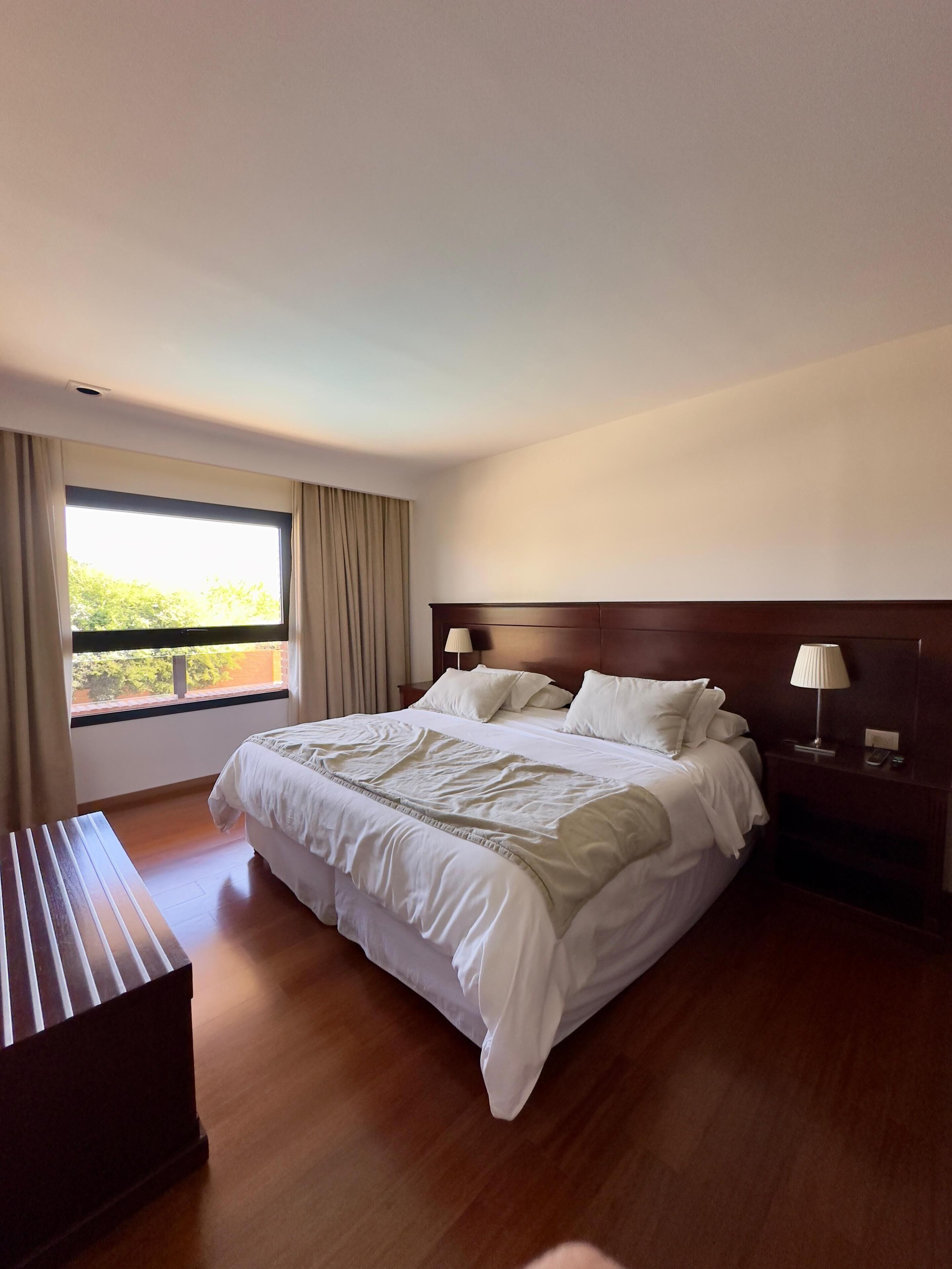 Una camera d'albergo con un letto grande, una panca di legno, un comodino con una lampada e una finestra con tende.
