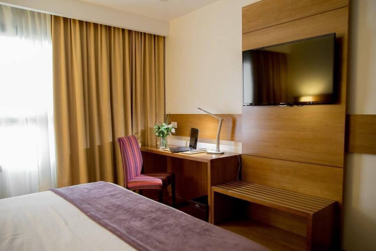 A hotel room with a bed, a desk with a laptop, a chair, a TV mounted on the wall, and a window with curtains.