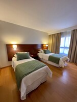 Quarto de hotel com duas camas, cada uma com um cobertor verde, cabeceira de madeira e luminárias laterais.