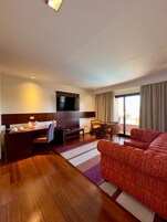 Quarto de hotel com chão de madeira, televisão presa à parede, uma secretária com cadeira, um sofá e uma mesa de jantar com cadeiras.