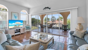 Ein geräumiges Wohnzimmer mit großem Fernseher, bequemen Sitzgelegenheiten und einem Balkon mit Blick auf Palmen und ein Schwimmbad.