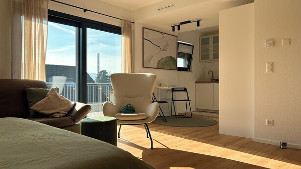 Una moderna camera d'albergo con un letto grande, un angolo salotto con una poltrona e poggiapiedi, una kitchenette e un balcone con vista su un edificio lontano.