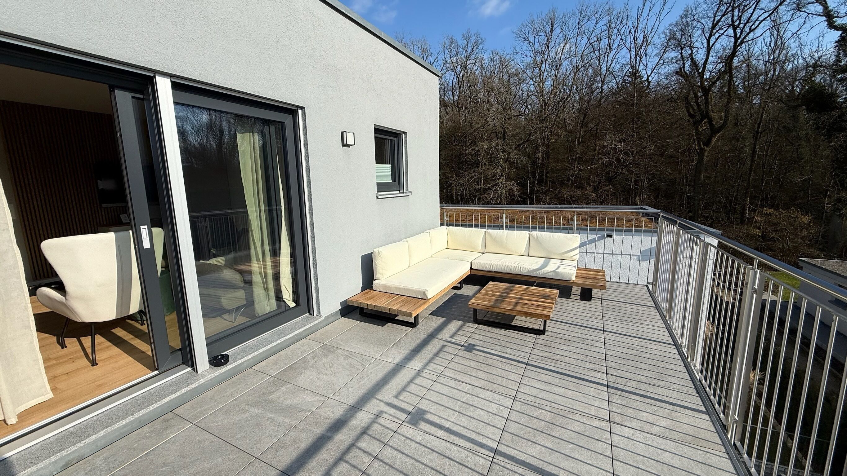 Un balcon extérieur moderne, doté d’un canapé et d’une table en bois, d’une porte vitrée donnant sur un espace intérieur et d’une rambarde en métal.