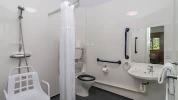 A bathroom with a shower, toilet, and sink. There is a white chair and a handrail.