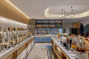 Un espace buffet comprenant différents postes de service pour les aliments, un comptoir central où les plats sont présentés, et un espace repas avec des tables et des chaises.