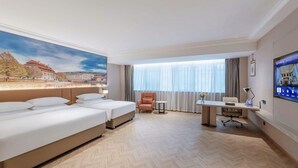 Ein Hotelzimmer mit zwei Betten, einem Schreibtisch, einem Sessel, einem Fernseher und einem großen Fenster mit Vorhängen.