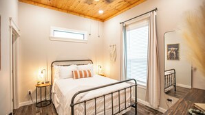 Een slaapkamer met een houten plafond, een bed met metalen frame, nachtlampjes, een raam met gordijnen en een klein nachtkastje.