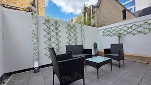 An outdoor patio with wicker furniture and a glass table, surrounded by a white wall with climbing plants. - City Home Paddington by Kravens (London)