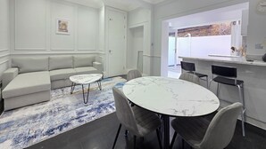 A modern living room with a sofa, a round table, and chairs. There is a kitchen area with a counter and a window. - City Home Paddington by Kravens (London)