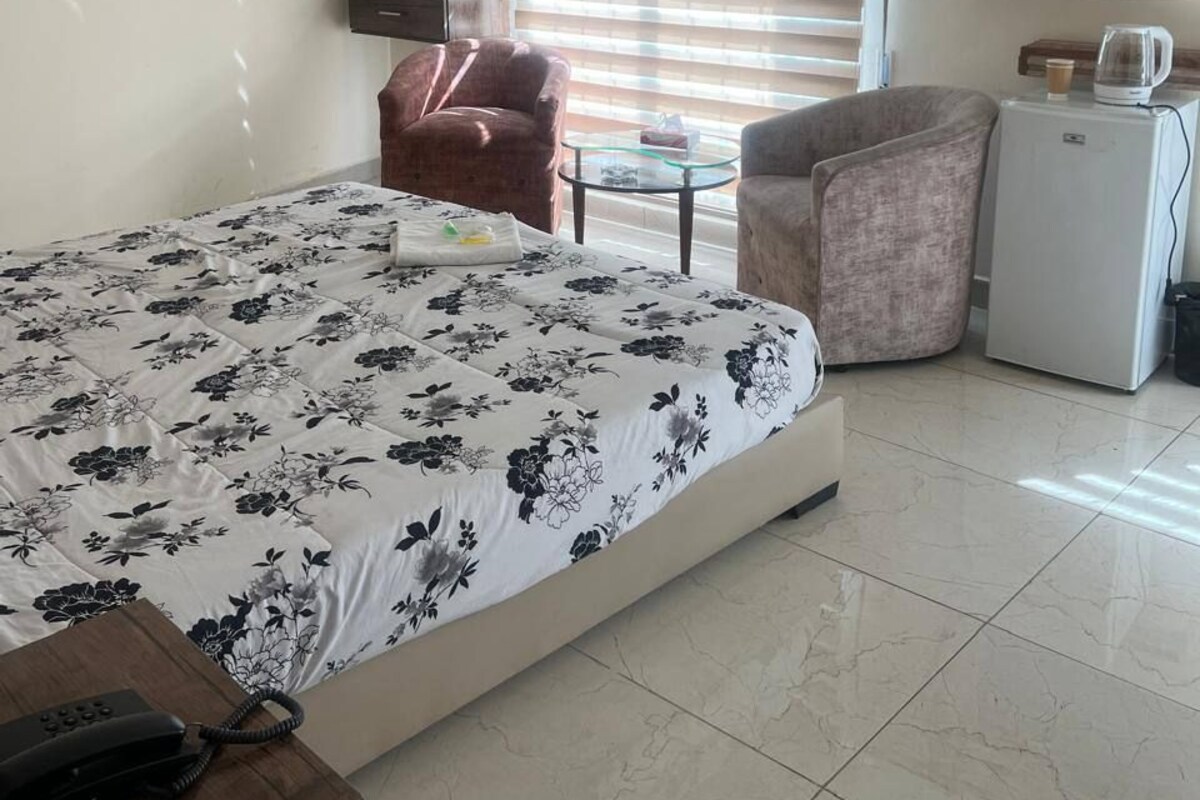 A hotel room with a bed, a desk with a phone, a chair, a small table with a cup, a TV, and a refrigerator.