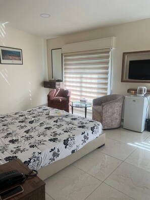 A hotel room with a bed, a desk with a phone, a chair, a small table with a cup, a TV, and a refrigerator. - Larnaka Hotel (Amman)