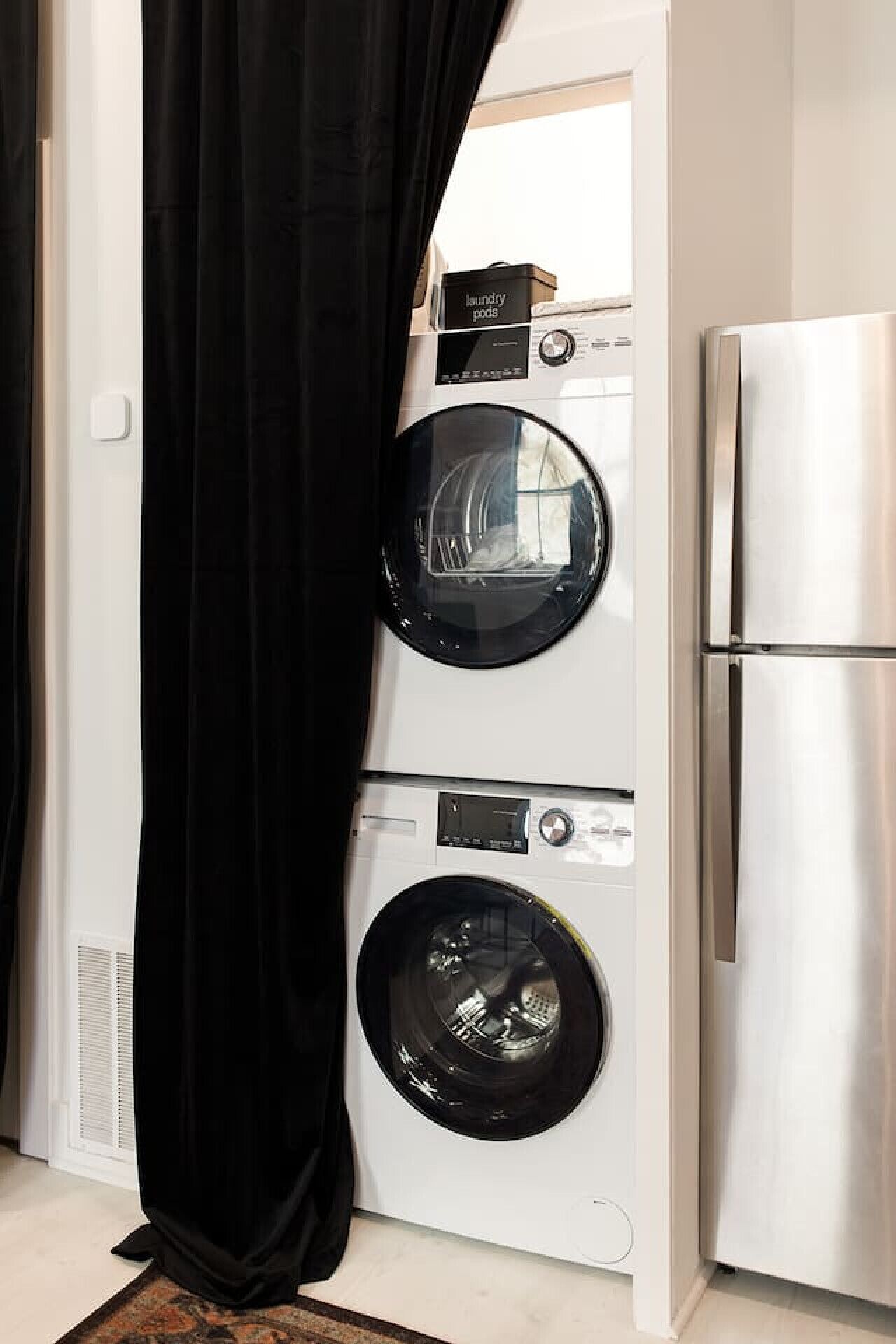 A compact laundry room with a front-loading washer and dryer, a stainless steel refrigerator, and a black curtain.