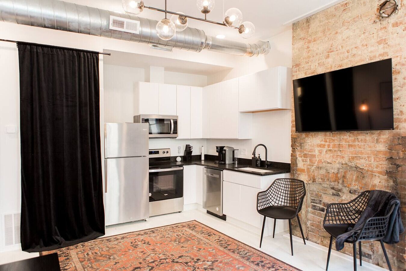 A compact kitchen with white cabinets, stainless steel appliances, a black countertop, and a black chair.