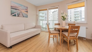 Un salon avec un canapé beige, une table à manger en bois avec des chaises, un vase de fleurs et un tableau représentant une jetée.