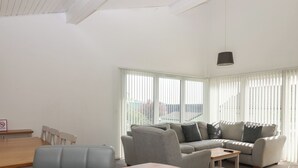 Een moderne woonkamer met een houten eettafel, een grijze bank en een groot raam met zonwering.
