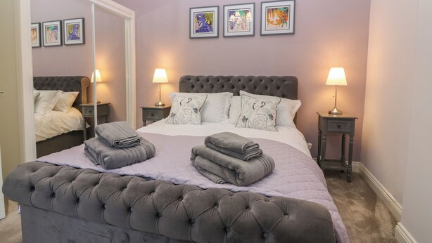 A neatly made bed with a tufted headboard, two bedside tables with lamps, and three framed artworks on the wall. - 60E Wellgate (Lanark)