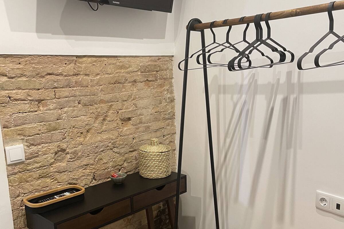 Une pièce avec un mur en briques, un téléviseur à écran plat fixé au-dessus d’un banc en bois, une petite table avec un panier et un porte-vêtements.