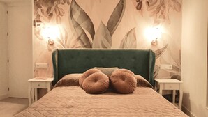Une chambre à coucher avec un grand lit, une tête de lit recouverte d’un tissu vert et un papier peint à motifs floraux. On y trouve deux tables de chevet avec des lampes.
