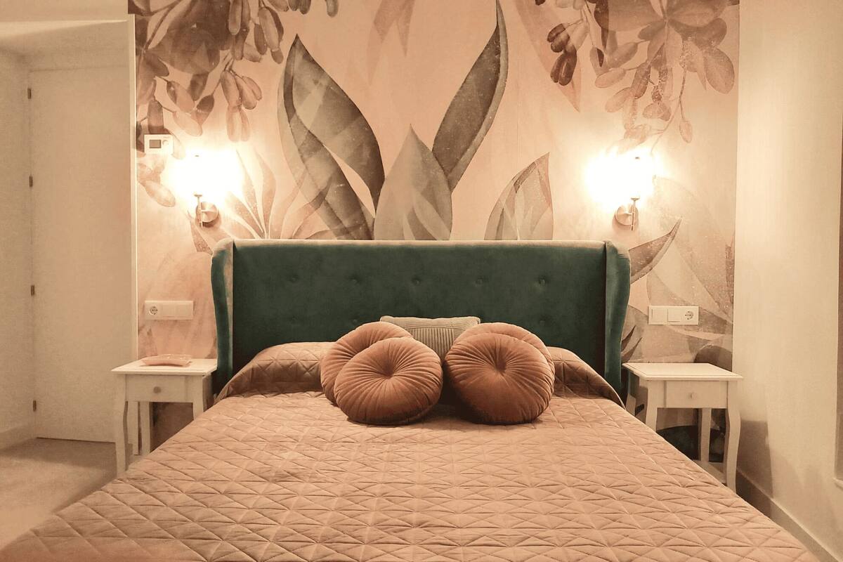 Une chambre à coucher avec un grand lit, une tête de lit recouverte d’un tissu vert et un papier peint à motifs floraux. On y trouve deux tables de chevet avec des lampes.