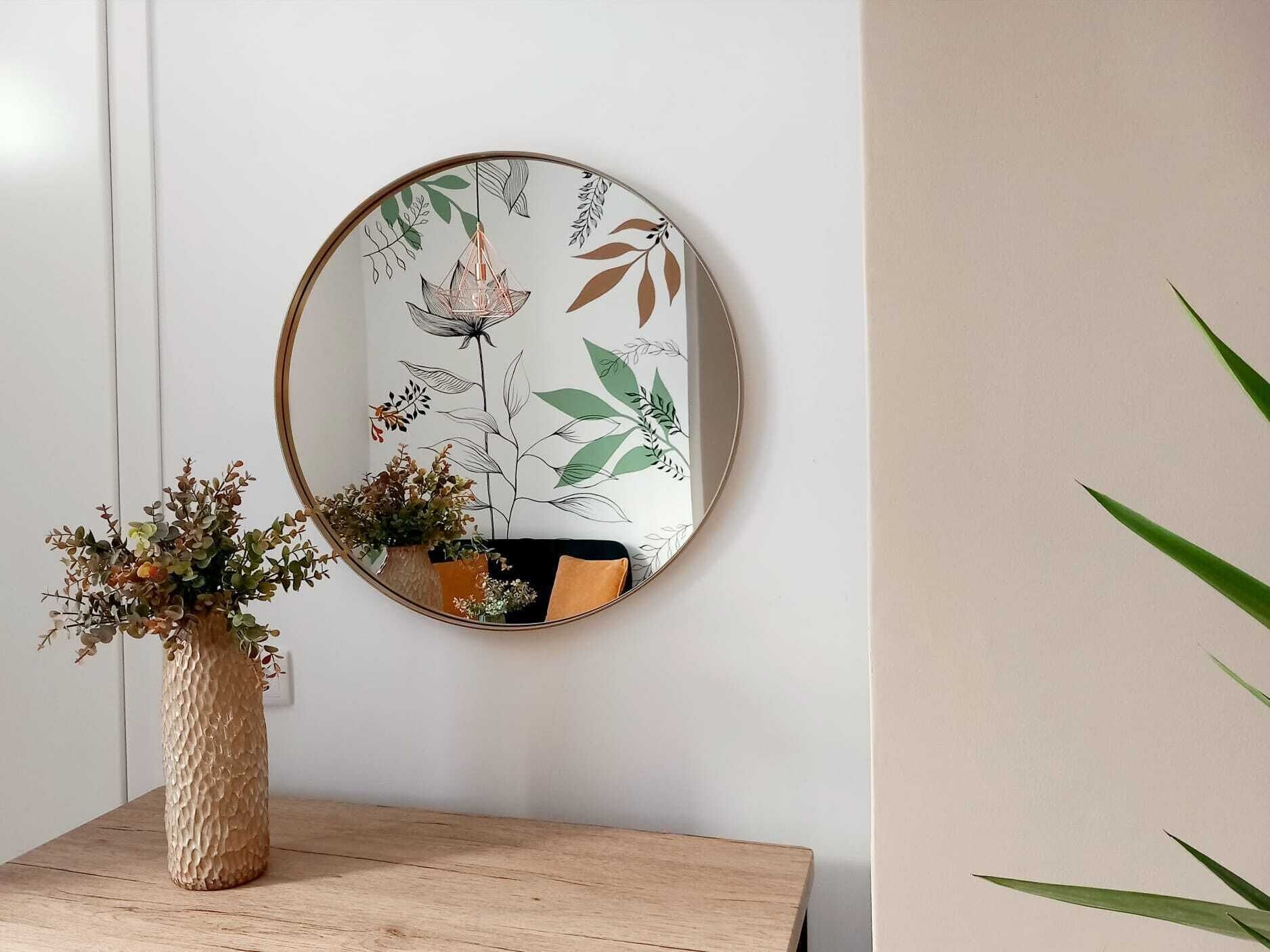 Espejo redondo con diseño floral en una pared, una mesa de madera con un jarrón de flores secas y una planta en maceta.