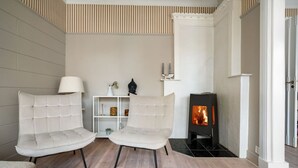 A modern living room with two beige armchairs, a white lamp, a small shelf with decorative items, and a black wood-burning stove. - Experience the luxury lifestyle 100m from Bryggen! (Bergenhus)