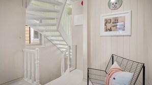 A staircase with white railings leading to a second floor, a framed collage on the wall, and a black wireframe chair with pillows.