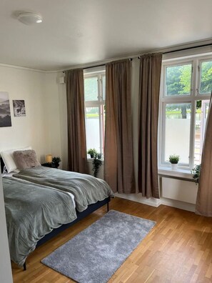 A bedroom with a bed, a grey rug, wooden flooring, a window with curtains, and a small plant on the windowsill.