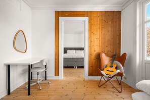 Een kamer met een wit bureau, een grijze bureaustoel, een gitaar in een lederen fauteuil, een bed met witte lakens, en een houten wand.