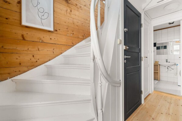 Een trap met witte treden die leidt naar een donkere deur, een houten muur met een lijstwerk en een badkamer met witte tegels en een wastafel.