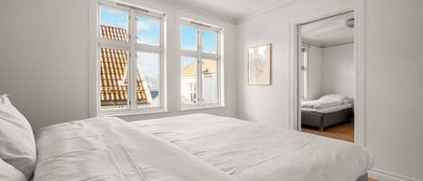 Quarto com uma cama grande, duas janelas, uma porta que dá para outro cômodo e um quadro na parede.