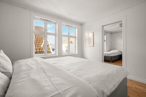 A bedroom with a large bed, two windows, a door leading to another room, and a framed picture on the wall.