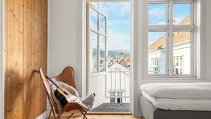 A bedroom with a bed, a wooden wall, a chair with a guitar, and a large window with a view of buildings. - Bryggen  Historical Townhouse I Rooftop Views! (Bergenhus)