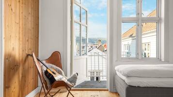 A bedroom with a bed, a wooden wall, a chair with a guitar, and a large window with a view of buildings.