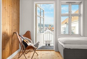 A bedroom with a bed, a wooden wall, a chair with a guitar, and a large window with a view of buildings.