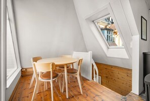 Een eetgedeelte met een houten tafel en stoelen, een raam met uitzicht op een gebouw, en een witte muur.