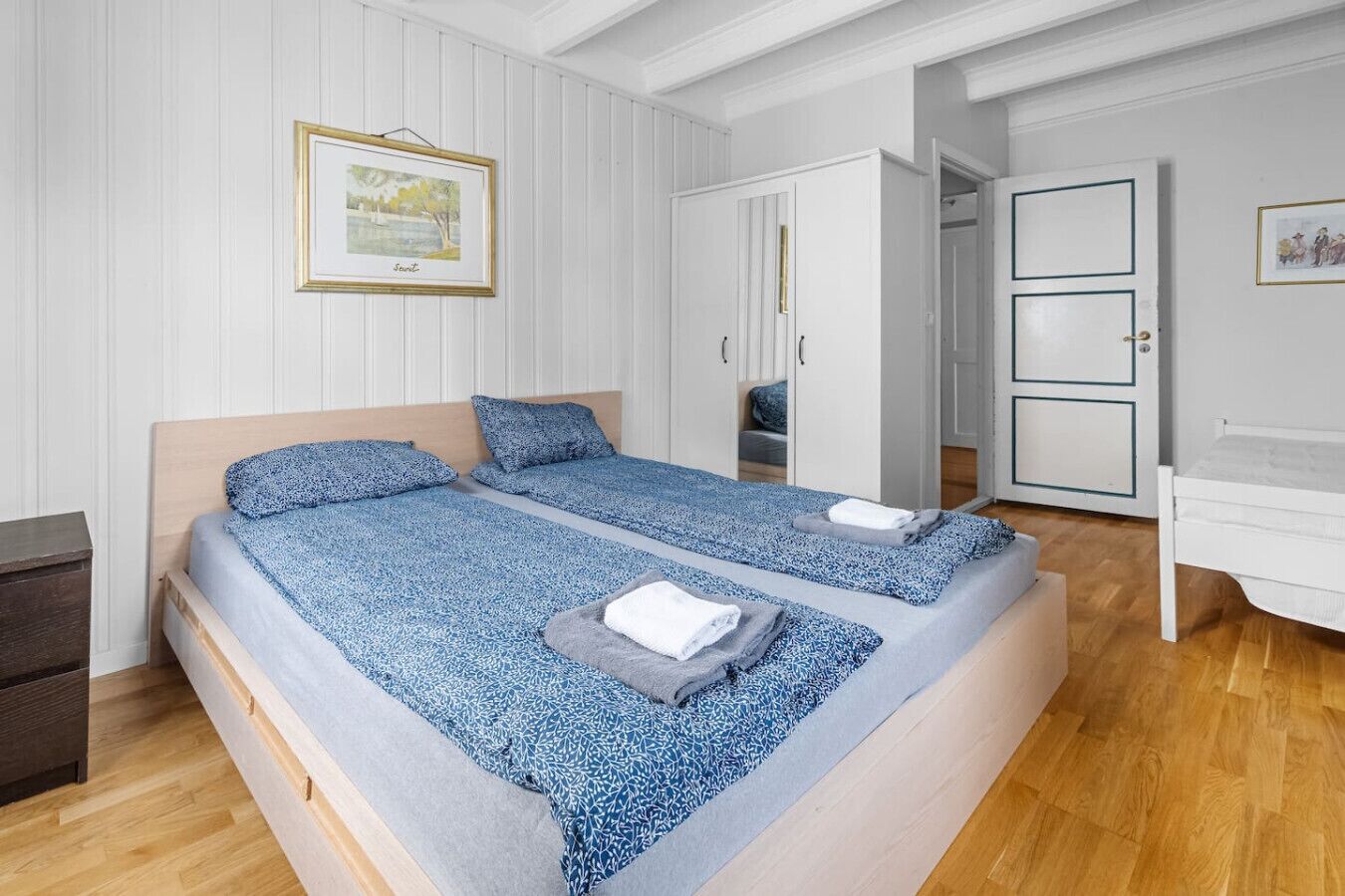 A double bed with a blue patterned bedspread, a wooden headboard, and a small bedside table. There is a wardrobe with a mirror and a framed picture on the wall.