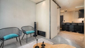 A modern room with two black wireframe chairs with teal cushions, a wooden coffee table, and a white rug. A tall black floor lamp is beside the chairs. The room has a minimalist design with a large window and a kitchen area visible in the background. - The Double Date I  Architectural (Bergenhus)