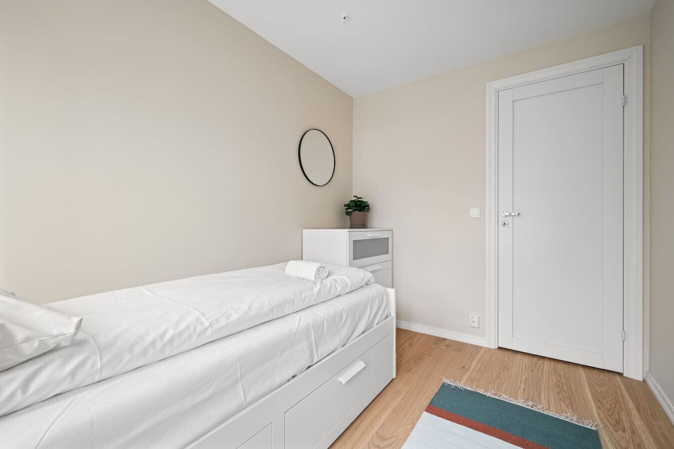 Una camera da letto con un letto, un comodino con una pianta, una porta e un piccolo tappeto.