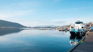 Un port tranquille où un ferry est amarré, une jetée qui s’avance dans l’eau et, au loin, un paysage vallonné.