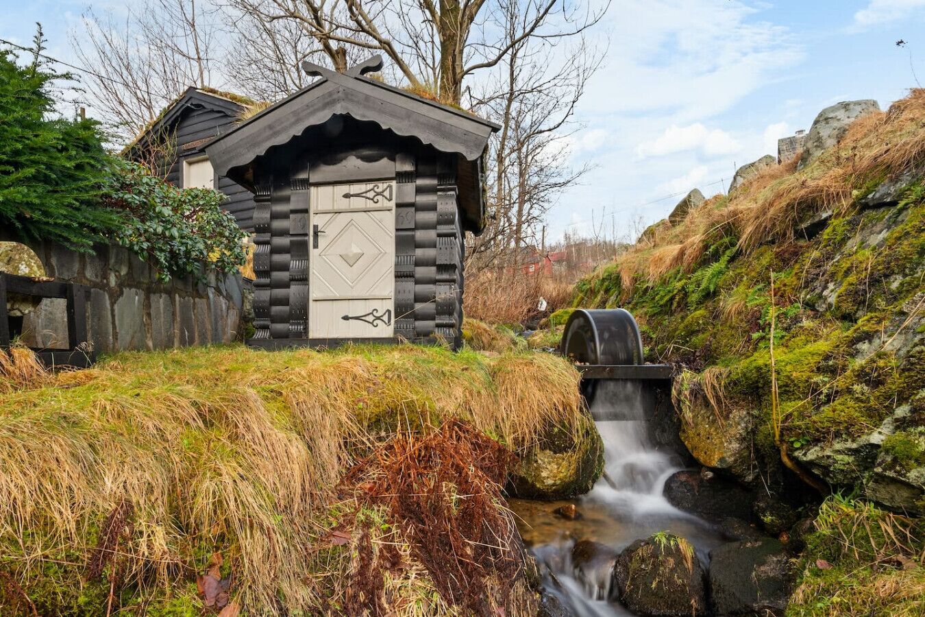 A rustic wooden cabin with a stone door, a small stream flowing through a rocky landscape, and a wooden fence surrounding the area.